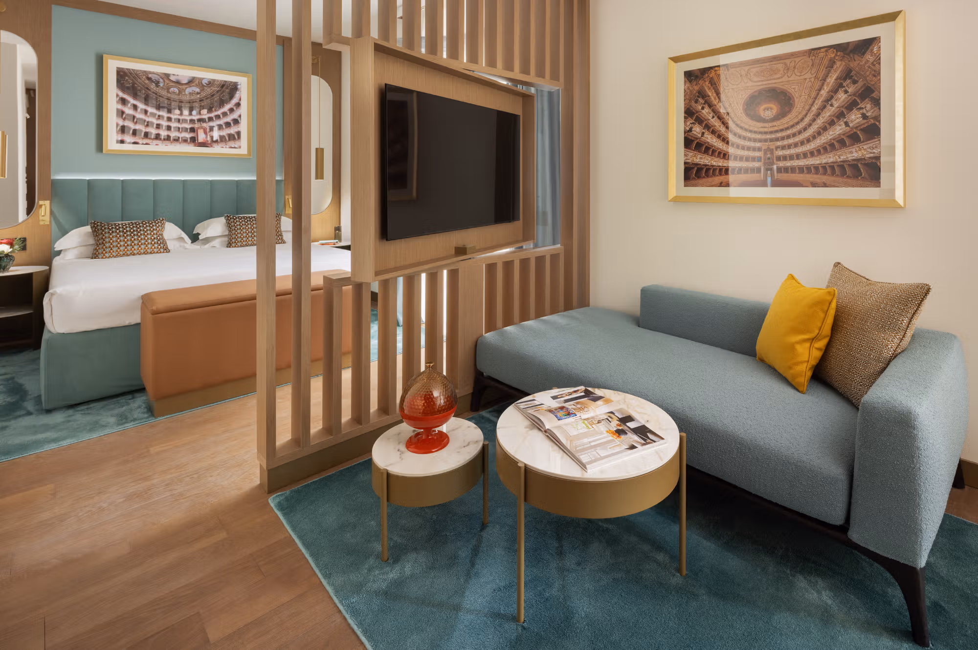 A studio flat at Teatro Luxury Apartments: a bed behind a wooden slat divider, a teal sofa and marble side tables, and framed prints of a theatre interior.